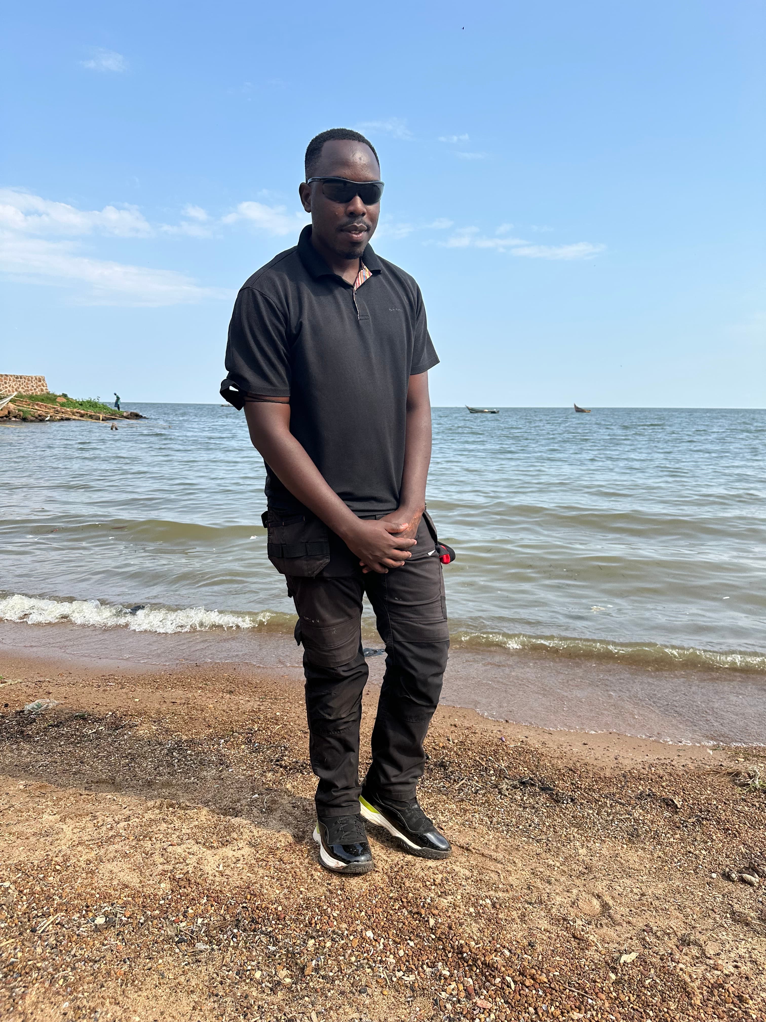 Man in black clothing and sunglasses standing on a pebbly beach by the water.