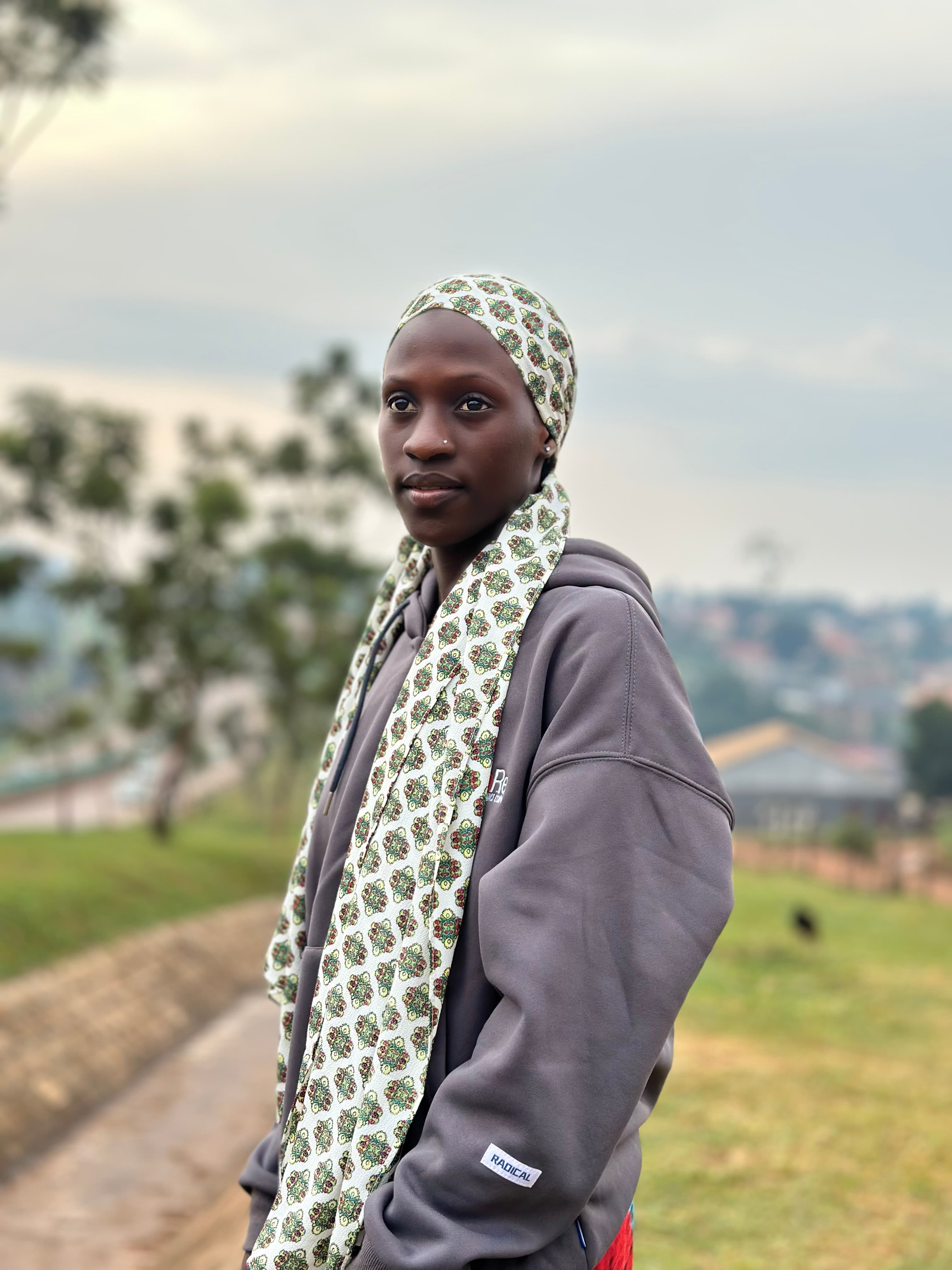 Young Black woman wearing a patterned headscarf and grey hoodie against a blurred landscape.