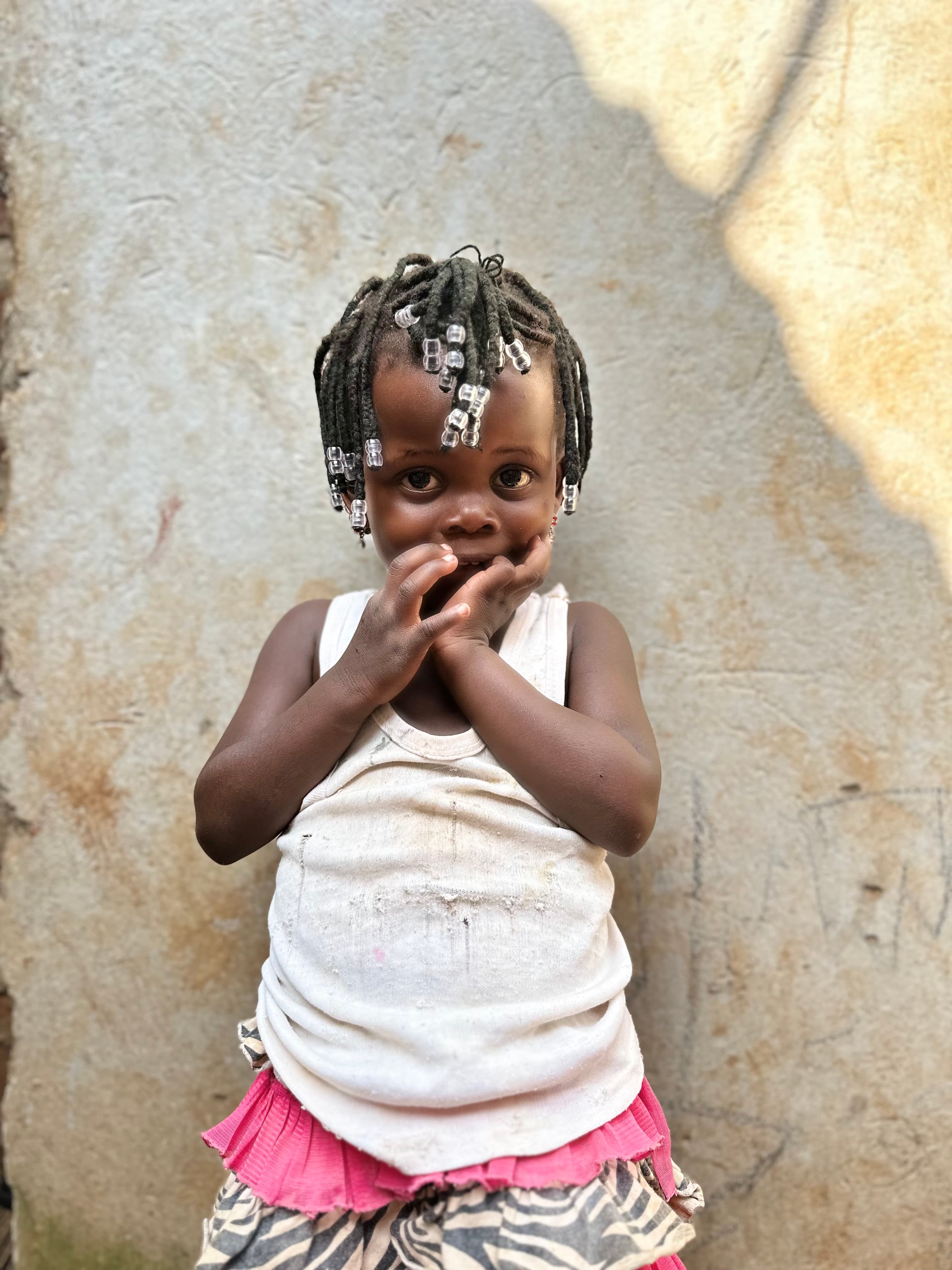 Shy young girl with beaded braids posing with hands to her mouth against a wall.