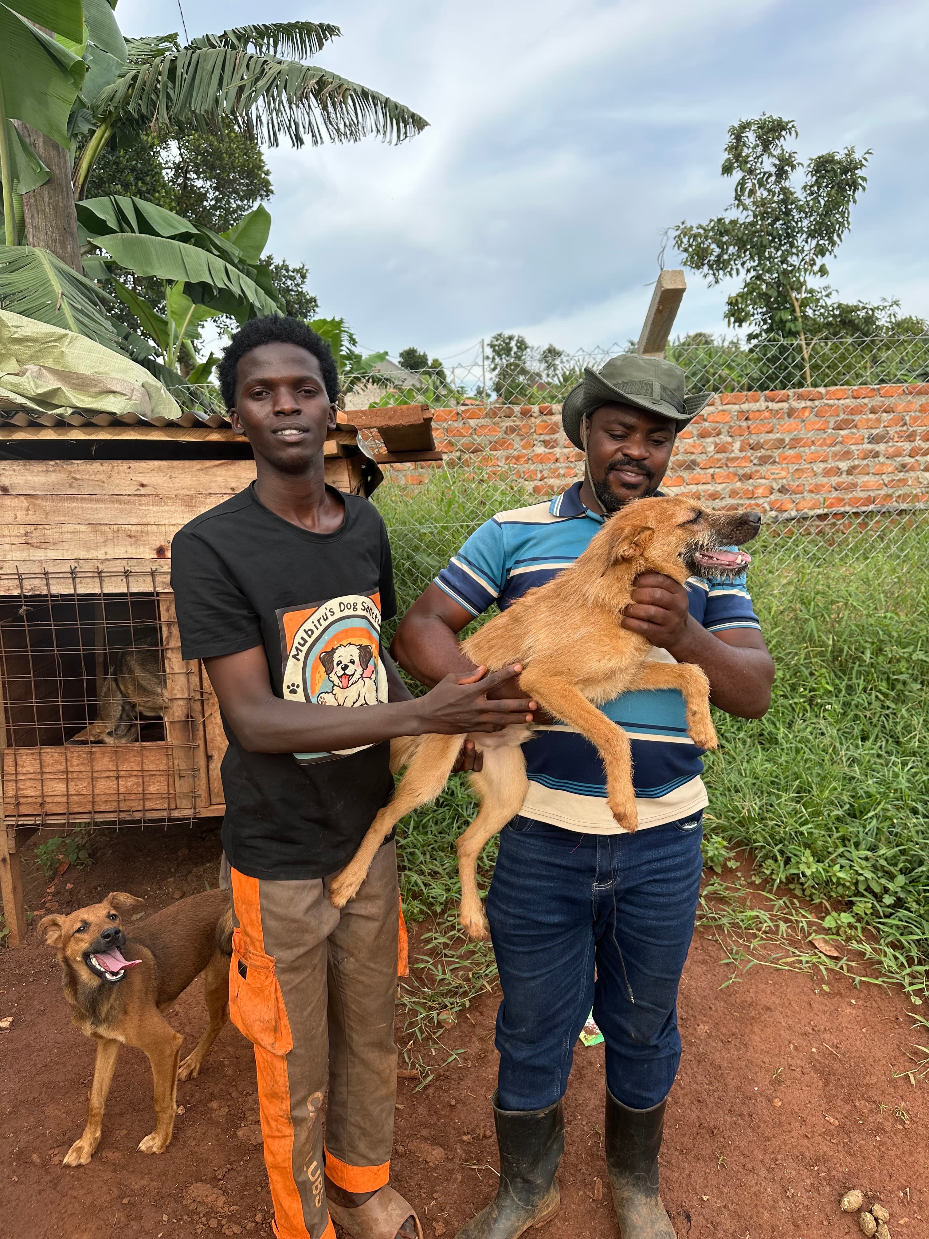 Two men holding a tan dog outdoors near a wooden kennel and another dog.