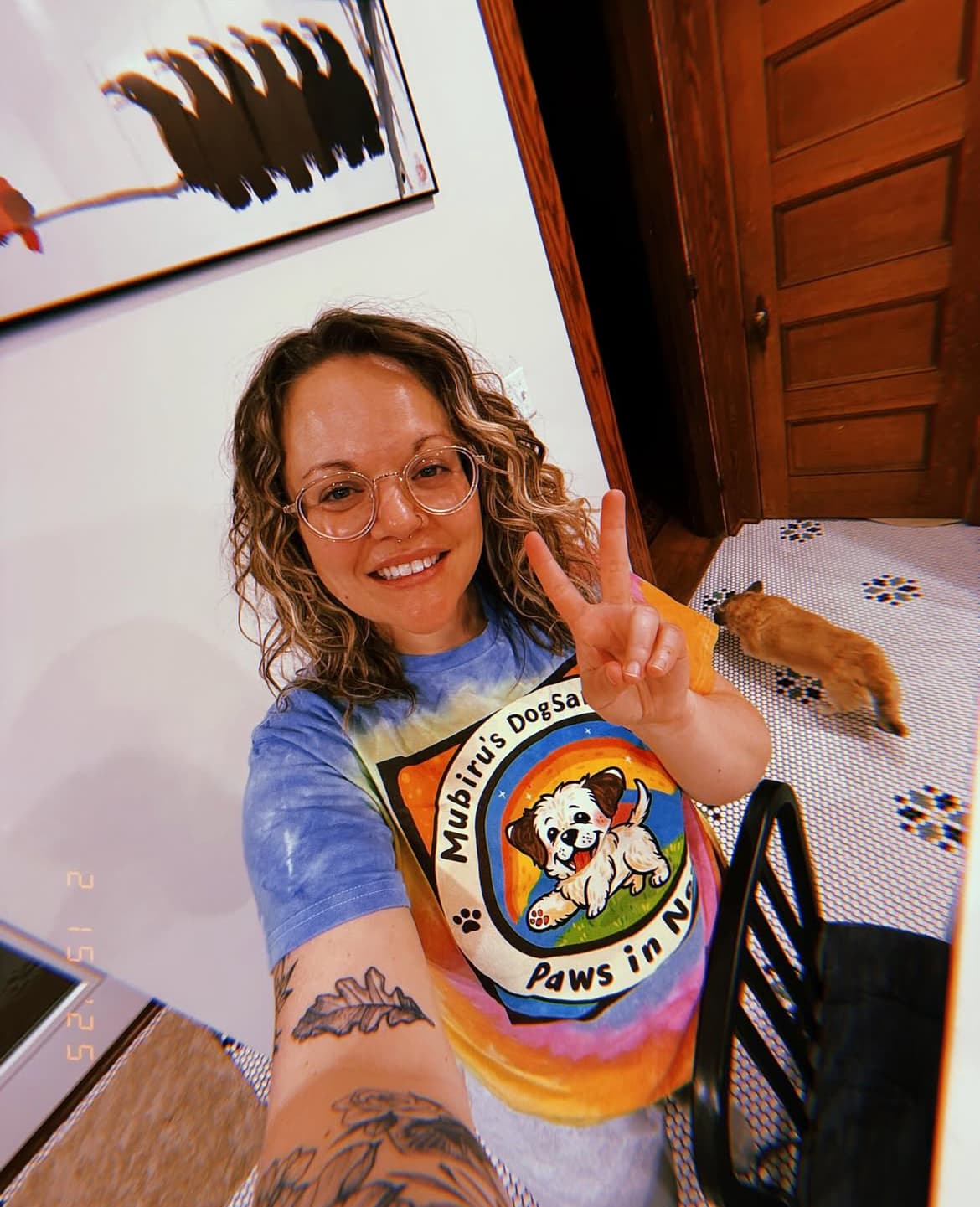 Smiling woman with curly hair and glasses in a tie-dye shirt, making peace sign.